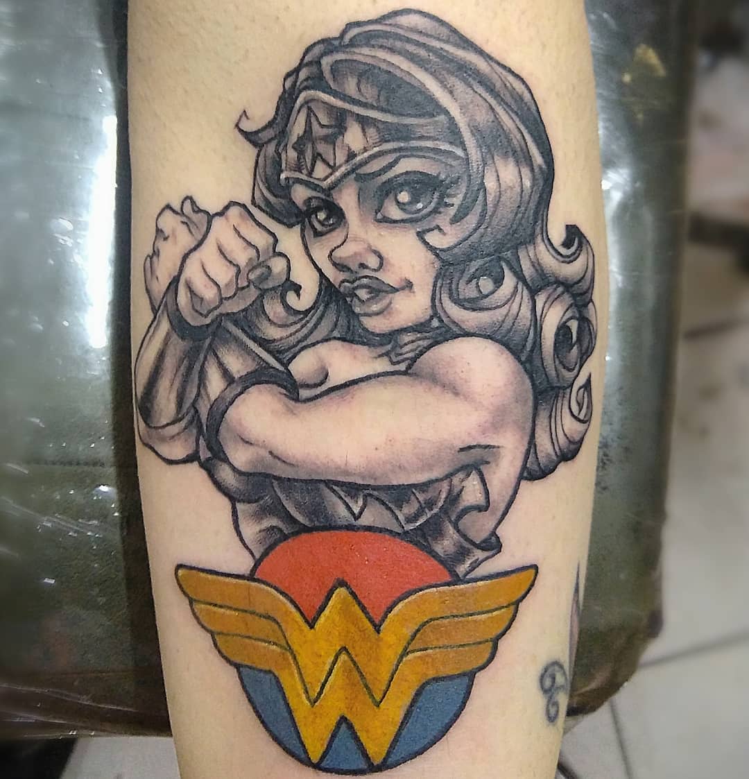 New school tattoo of Wonder Woman in grayscale with bold features, crossing arms over a colorful logo